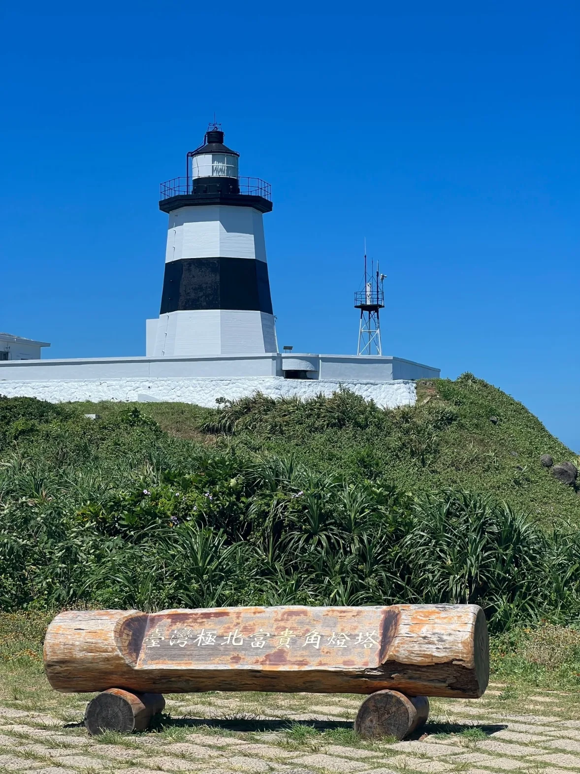The black and white banded Fuguei Cape lighthouse above windswept green scrub under a deep blue sky, a carved driftwood log sign in the foreground